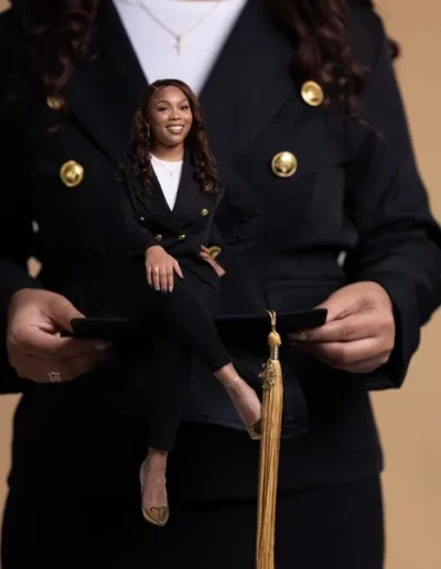 A woman in a black suit holds a graduation cap; a small version of herself appears to be sitting on the cap, smiling confidently.
