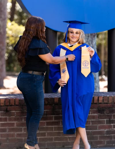 A person in a blue graduation gown and cap stands while another adjusts their stole outdoors in front of a brick wall.