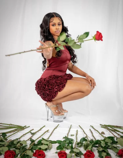 A woman in a red dress and clear heels squats on a white background, holding out a red rose. Several red roses are arranged on the floor in front of her.