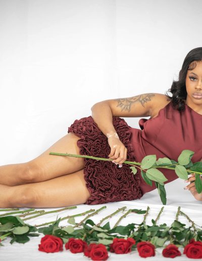 Woman in a burgundy dress lies on her side holding a red rose, surrounded by several roses arranged on a white surface.