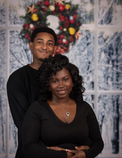 A man and woman stand closely in front of a frosted window backdrop with a holiday wreath, both wearing black tops and smiling at the camera.