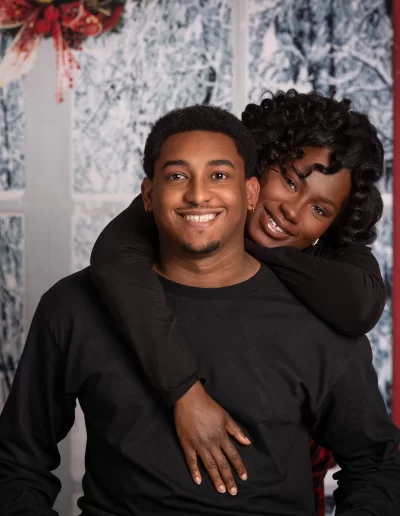 A smiling woman hugs a smiling man from behind in front of a winter-themed backdrop with a holiday wreath and red curtains.