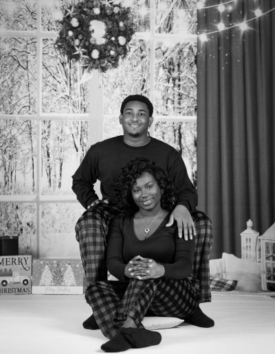 A man and woman in matching plaid pajamas pose indoors in front of a winter-themed backdrop, with holiday decorations visible in the background.