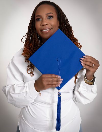 A woman in a white shirt holds a blue graduation cap and tassel, smiling at the camera against a neutral background.