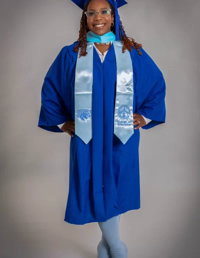 A person wearing a blue graduation cap and gown with a light blue stole stands smiling, facing the camera against a plain background.