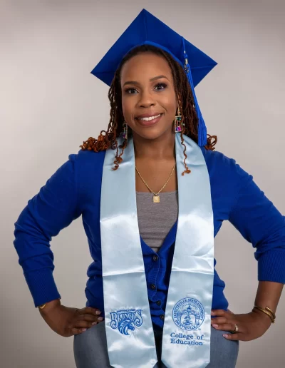 A person wearing a blue graduation cap, blue sweater, and a light blue stole with educational emblems poses and smiles in front of a plain background.