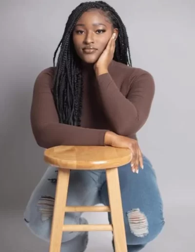 A woman with long braids sits on a wooden stool, resting her face on her hand. She wears a brown top and ripped blue jeans against a plain gray background.