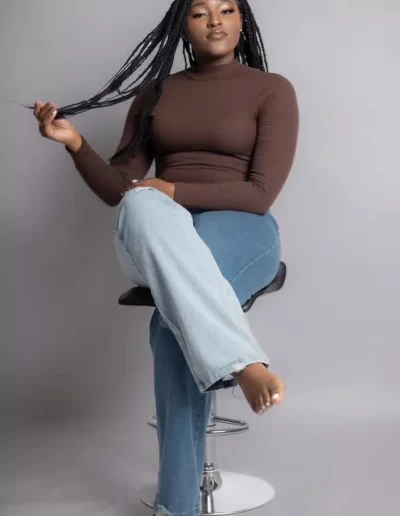 A woman with long braids sits barefoot on a stool, wearing a brown turtleneck and blue jeans, against a plain gray background.