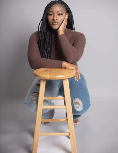 A woman with long braids wearing a brown top and ripped jeans sits on the floor, resting her arm and head on a wooden stool, against a plain gray background.