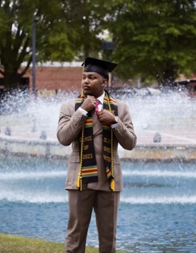 A graduate in a tan suit, kente stole, and cap stands in front of a fountain on campus with trees and buildings in the background.