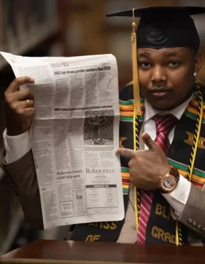 A man in graduation attire sits in a library, holding and pointing at a newspaper.