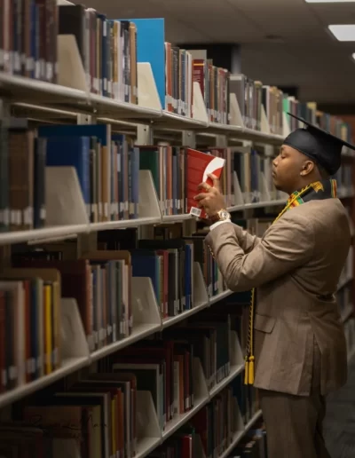 A person wearing a graduation cap and stole selects a book from a shelf in a library.