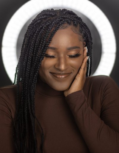 A woman with long braids and a brown top smiles with her eyes closed, one hand resting on her face, standing in front of a circular light.