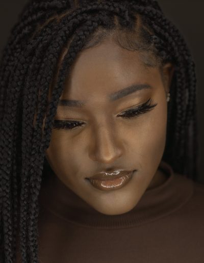 A woman with braided hair looks downward. She has glossy lips, long eyelashes, and is wearing a brown top against a dark background.