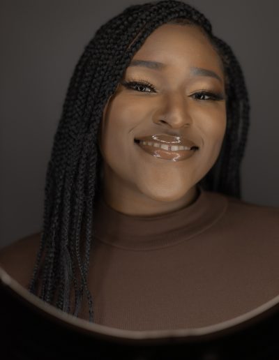 A woman with long braided hair, wearing a brown top, smiles at the camera against a neutral background.
