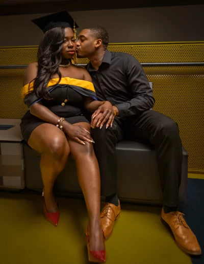 A woman in a graduation cap and dress sits next to a man in black attire; he kisses her on the cheek as they hold hands.