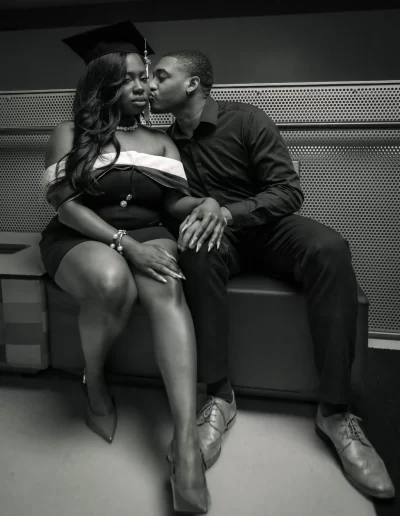 A woman in a graduation cap and off-shoulder dress sits next to a man in dark clothing who is kissing her on the cheek. They are holding hands and sitting on a bench indoors.