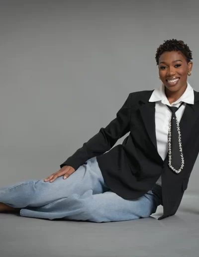 A person sits barefoot on the floor against a gray background, wearing a black blazer, white shirt, tie, and light blue jeans, smiling at the camera.