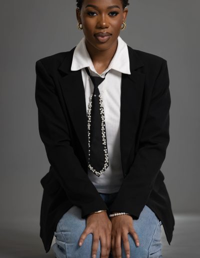 A person with short hair in a black blazer, white shirt, patterned tie, and jeans kneels on the floor against a gray background.