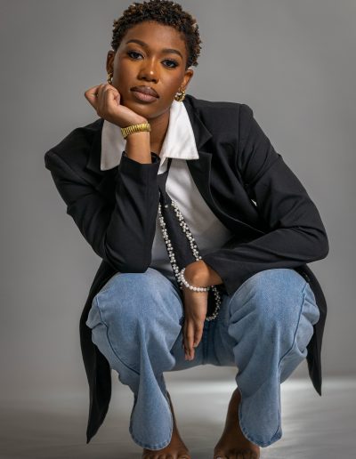 A person in a black blazer, white shirt, and blue jeans squats barefoot on the floor, resting their chin on their hand, against a plain grey background.