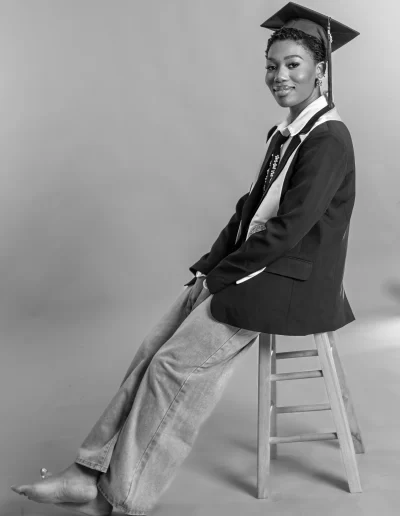 A person wearing a graduation cap, gown, and casual jeans sits barefoot on a wooden stool against a plain background.