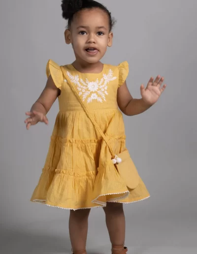 A young child in a yellow dress with white embroidery and a small matching purse stands against a plain gray background, looking forward with hands raised.