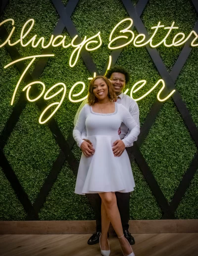 A smiling couple poses in front of a green wall with a neon sign that reads "Always Better Together." The woman wears a white dress and the man wears a white shirt and dark pants.