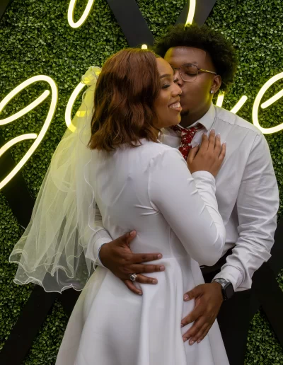 A couple poses in wedding attire; the person in a white dress and veil smiles as the other, in a white shirt and tie, kisses their cheek in front of a green wall with yellow neon writing.