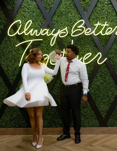 A woman in a white dress and heels dances with a man in a white shirt and black pants in front of a green wall with the words "Always Better Together" in neon.