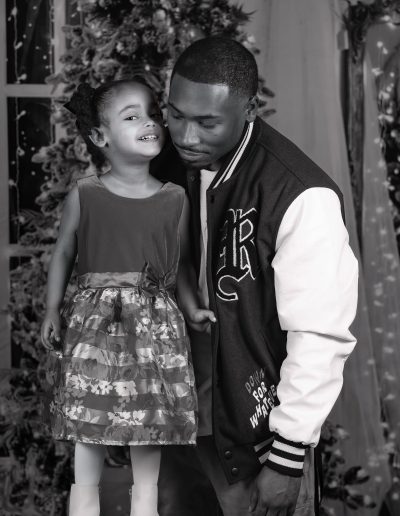 A young girl stands on a stool smiling beside an adult man in a varsity jacket, with a decorated Christmas tree in the background.