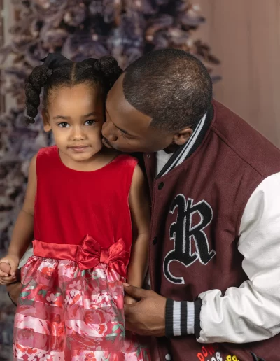 A man kisses a young girl on the cheek as she stands in front of a decorated tree, wearing a red dress with a floral skirt.